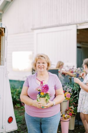 Person in pink shirt holding colorful flower arrangement outside white barn building with flower displays.