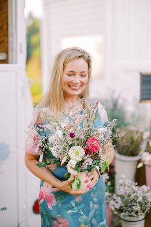 Someone wearing a vibrant floral dress smiles while holding a beautiful mixed bouquet of fresh flowers against a white wall.