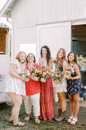 A group wearing summer dresses poses in front of a white barn while holding colorful floral bouquets.