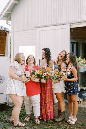 A group wearing colorful dresses and holding bouquets poses together in front of a white barn for a joyful celebration photo.