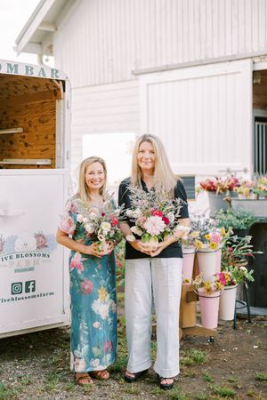 Two people stand together holding beautiful floral arrangements in front of a white barn with flower buckets nearby.