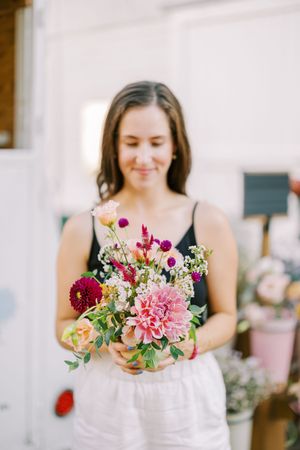 A person in white holds a vibrant bouquet with pink dahlias and mixed summer flowers against a light background.