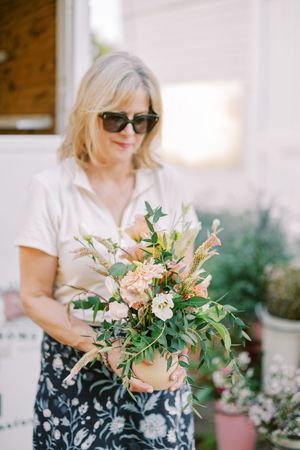 Person in sunglasses arranging a delicate peach and white floral bouquet outdoors.