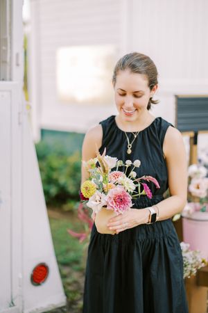 A person in a black dress carefully arranges delicate pink and white floral arrangements at an outdoor event.