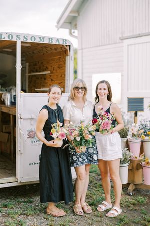 Three people stand in front of a vintage Possom Bay trailer holding beautiful floral bouquets at an outdoor market.