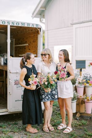 Group of friends shopping at vintage market stalls with pink floral arrangements and white barn walls in background.