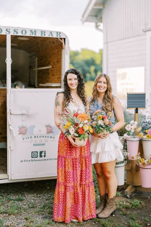 Two people stand together holding colorful bouquets in front of a vintage floral truck at a rustic outdoor event.