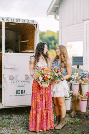 Two shoppers browse colorful bouquets and flowers outside a rustic white barn market with pink buckets.