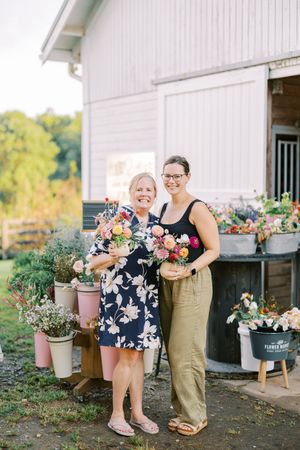 Customers shop for fresh flowers displayed in rustic buckets outside a white barn storefront.