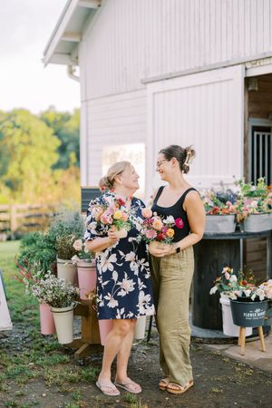 Two people share a joyful moment while surrounded by colorful potted flowers outside a white barn on a sunny day.