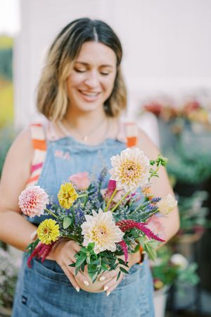 Person wearing denim overalls holding vibrant pink and yellow dahlia flower arrangements.