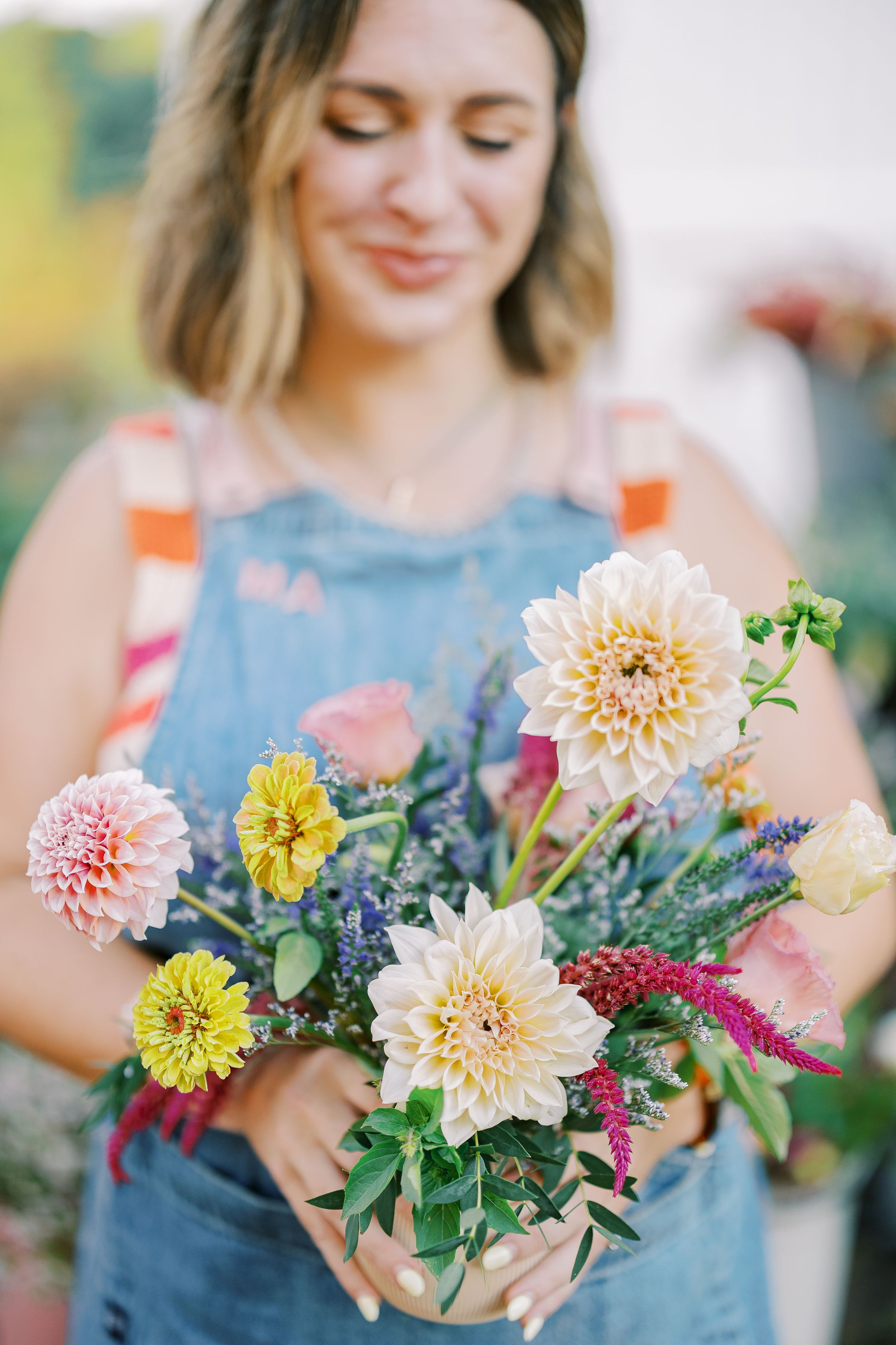 A bouquet of colorful dahlias and wildflowers held by someone wearing a blue denim overall dress in a sunny garden.