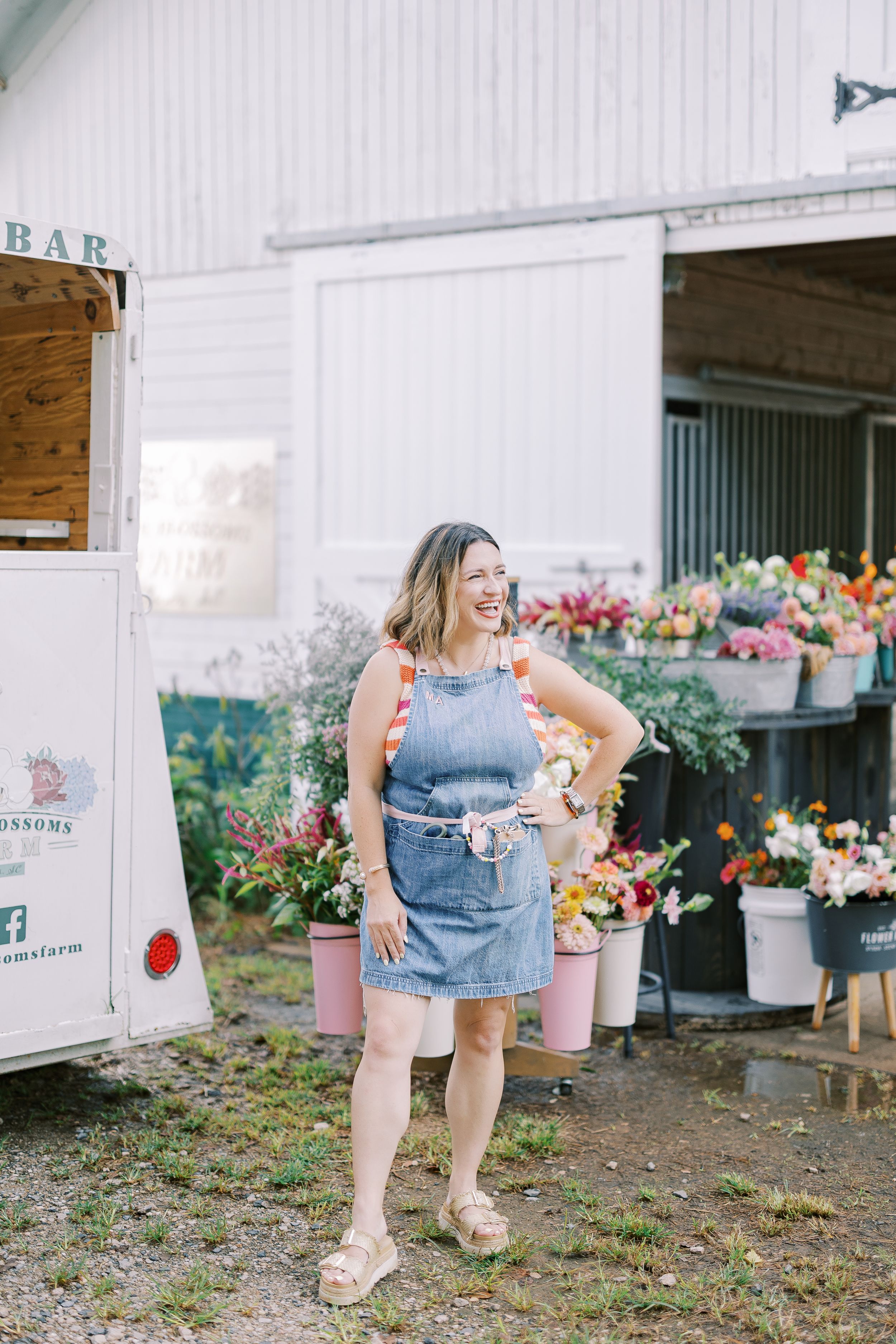 Person in denim dress stands outside rustic barn with flower buckets and garden displays.