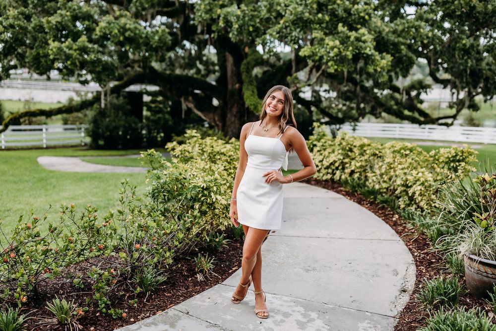 A toddler in brown boots walking along a garden path while wearing a white dress on a sunny day.