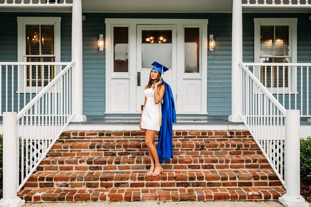 A graduate in blue cap and gown walks alongside a brick building with white stairs and columns.
