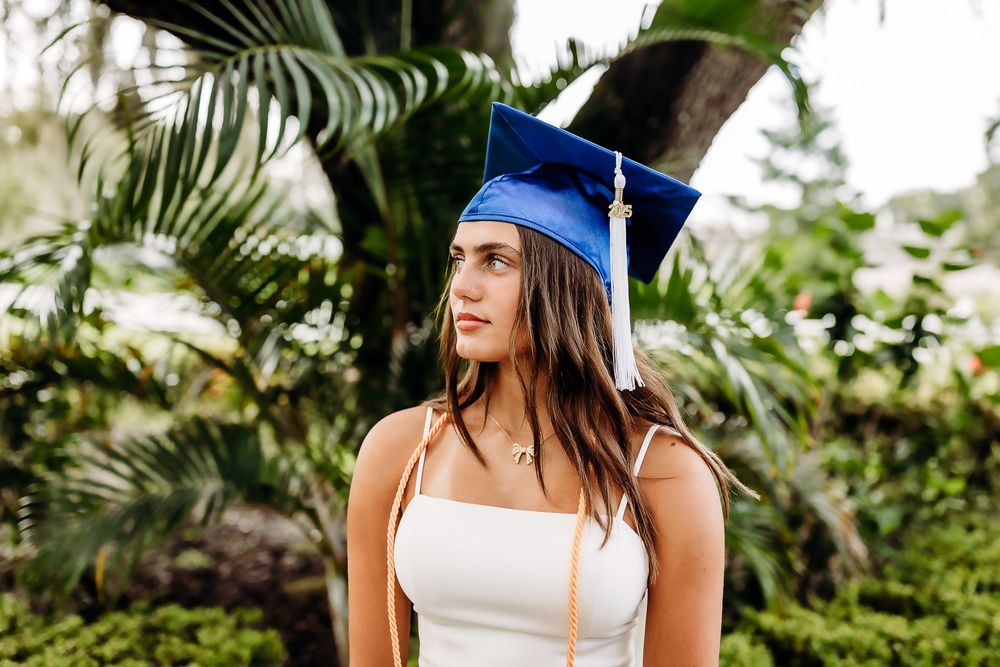 Graduate in blue cap and gown stands outdoors against lush tropical palm trees during commencement celebration.