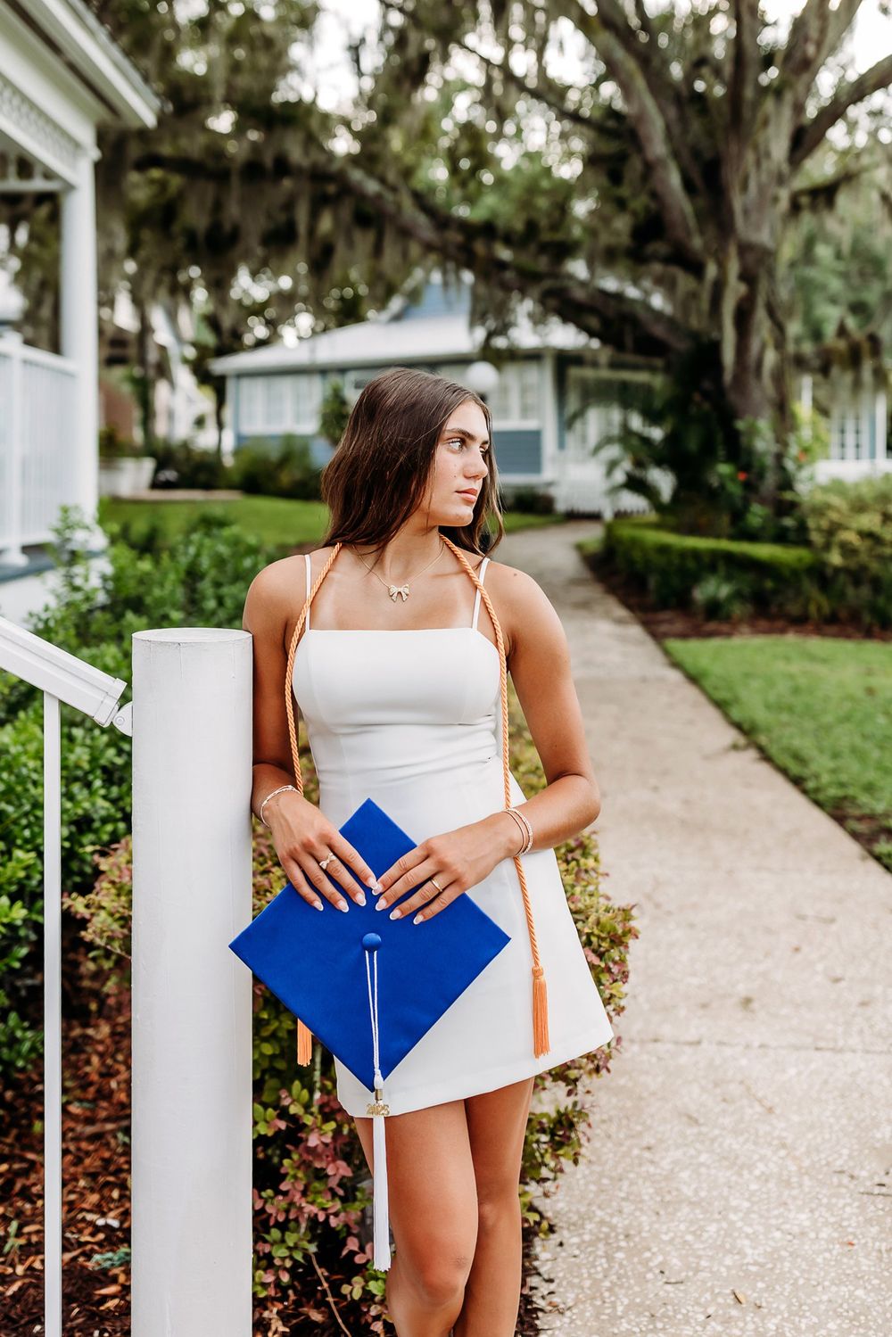 Graduate holds a blue diploma folder while walking along a scenic columned pathway surrounded by lush trees.