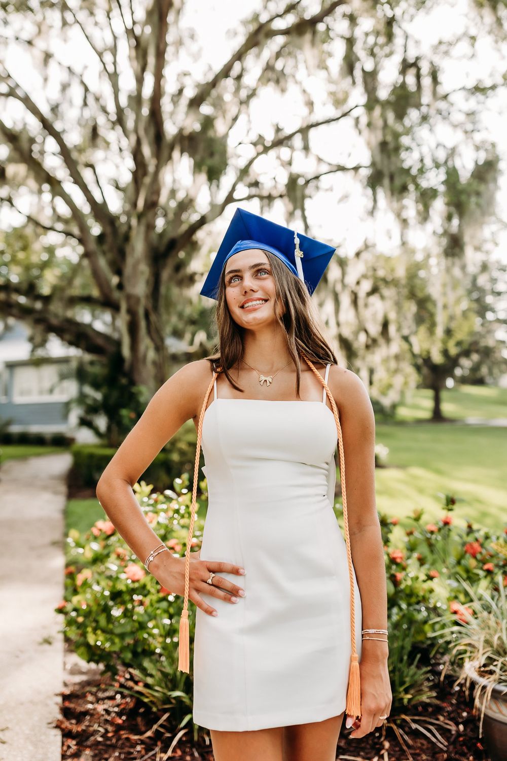 Graduate in blue cap and white dress stands in front of blooming garden under Spanish moss draped trees.