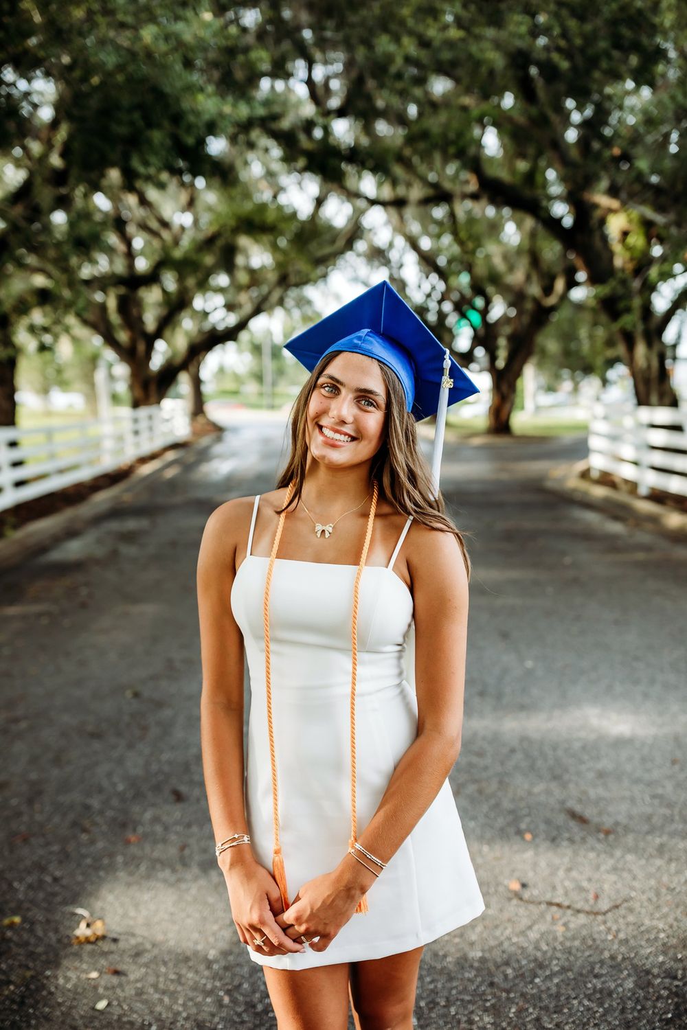 Graduate poses outside in a white strapless dress and blue cap with graduation stole under shaded tree-lined walkway.