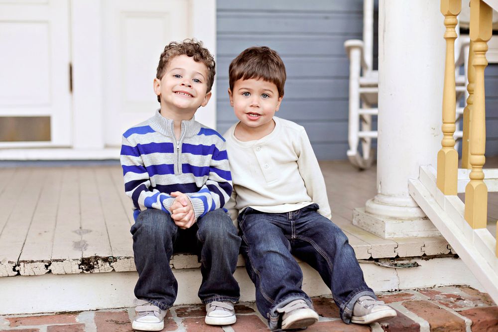 Two young children sit together on outdoor wooden steps wearing jeans and casual clothing.
