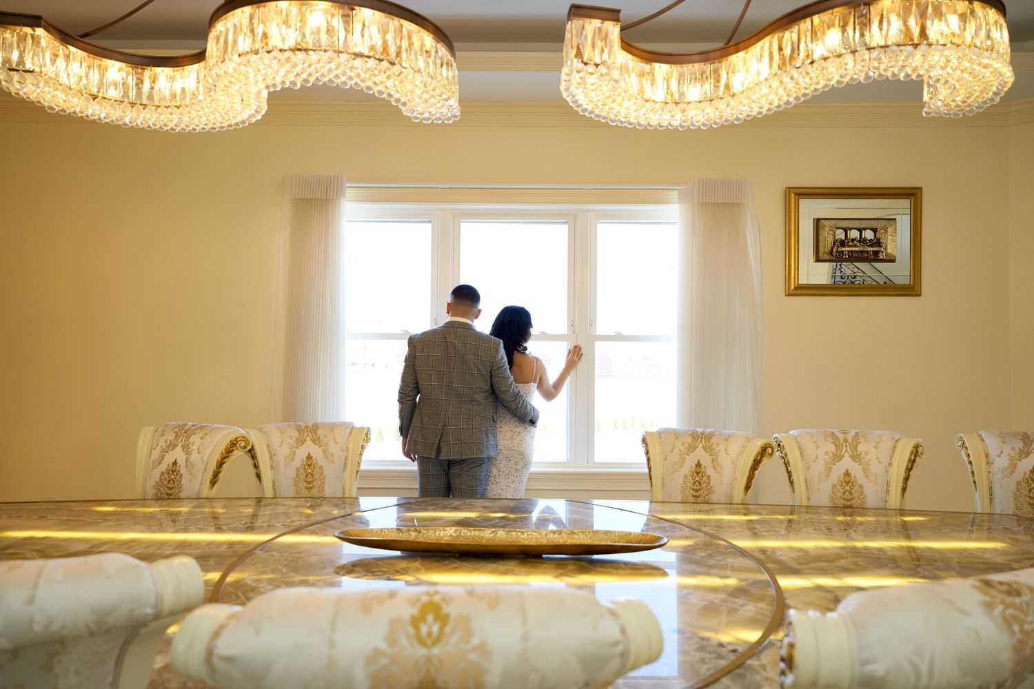Two figures stand by a window in an elegant banquet room with crystal chandeliers and gold-accented decor. Engagement photoshoot
