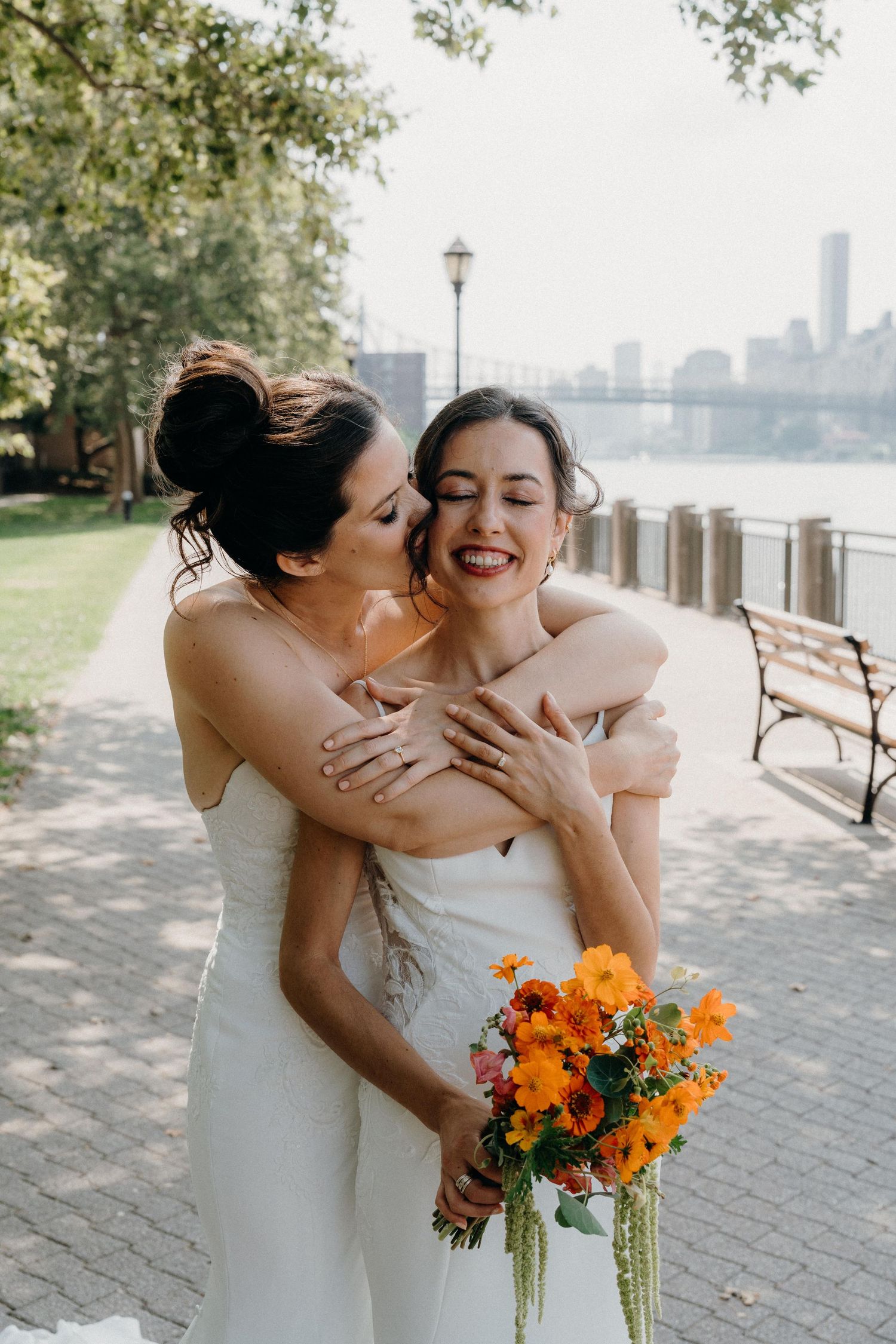 Two brides share a joyful embrace while holding a vibrant orange bouquet along a waterfront promenade.