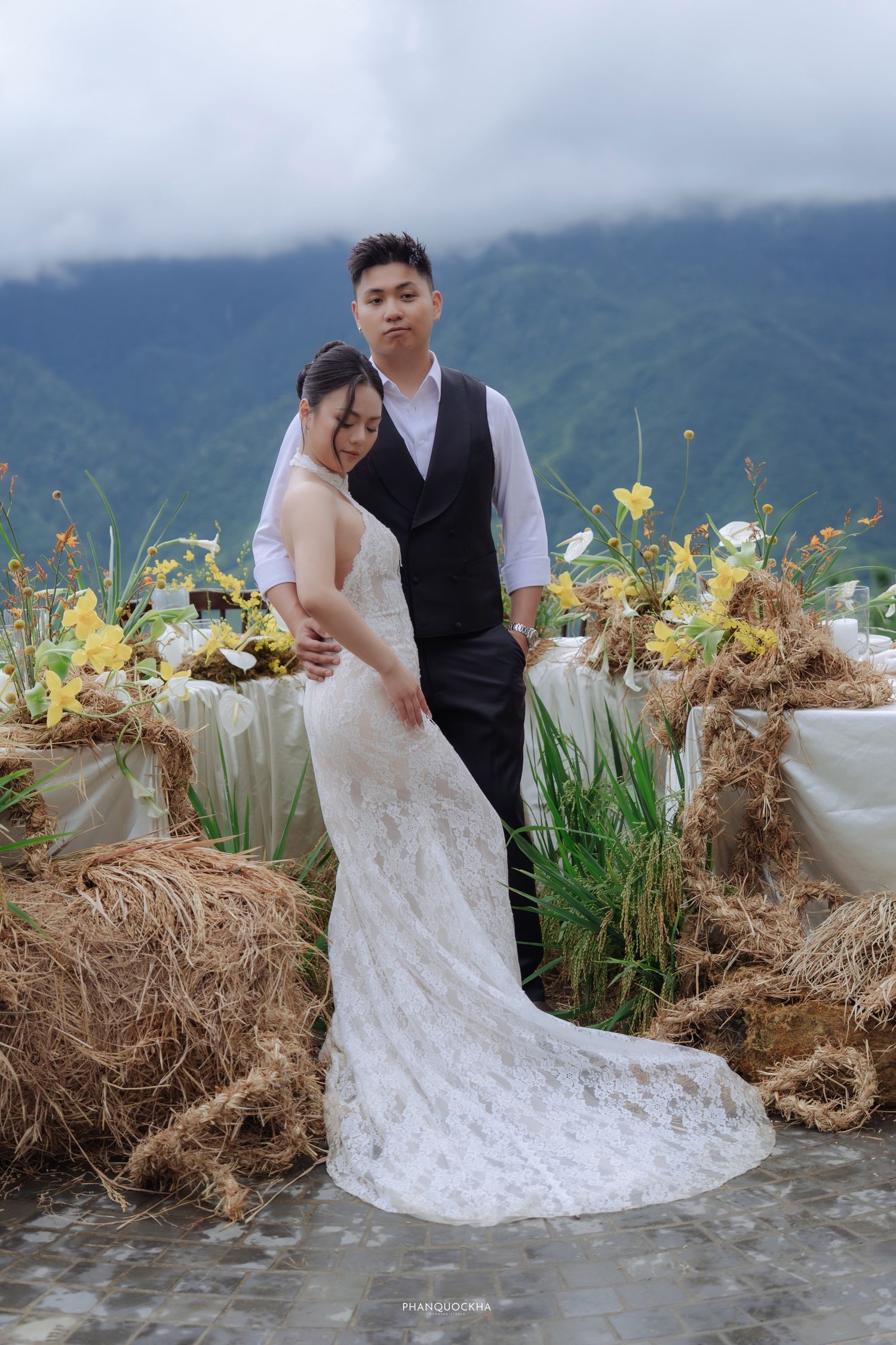 Bride in lace wedding gown and groom in black vest posing together before a mountain backdrop with yellow floral decor.