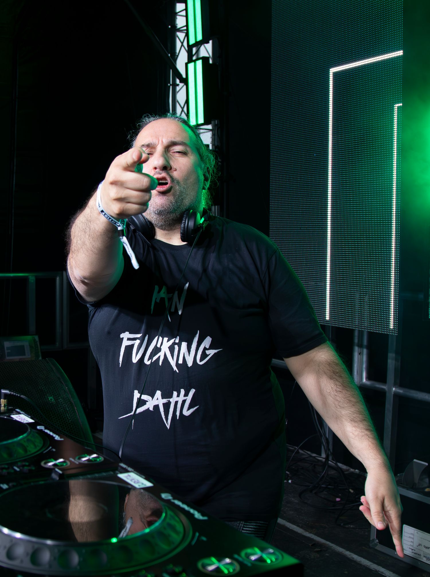 A performer in a black t-shirt and long hair energetically singing into a microphone on stage with green lighting effects.Ian Van Dahl
