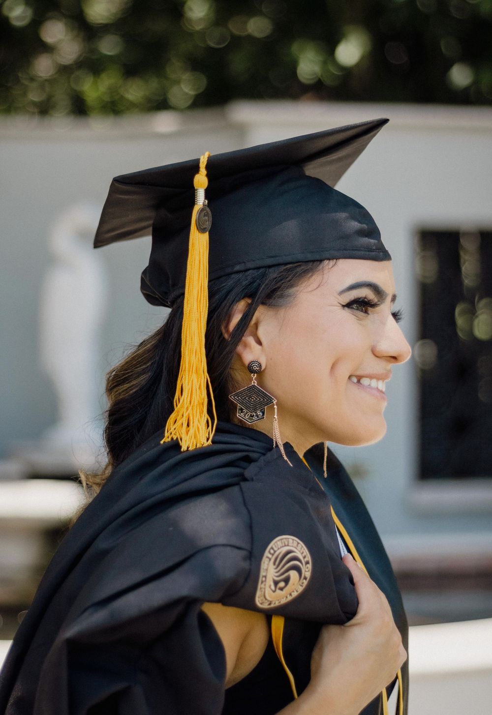 Smiling graduate in black cap and gown with gold tassel posing outdoors at graduation ceremony.