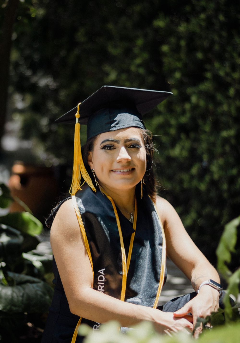 Female graduate smiling in black cap and gown with gold tassel surrounded by lush green foliage.