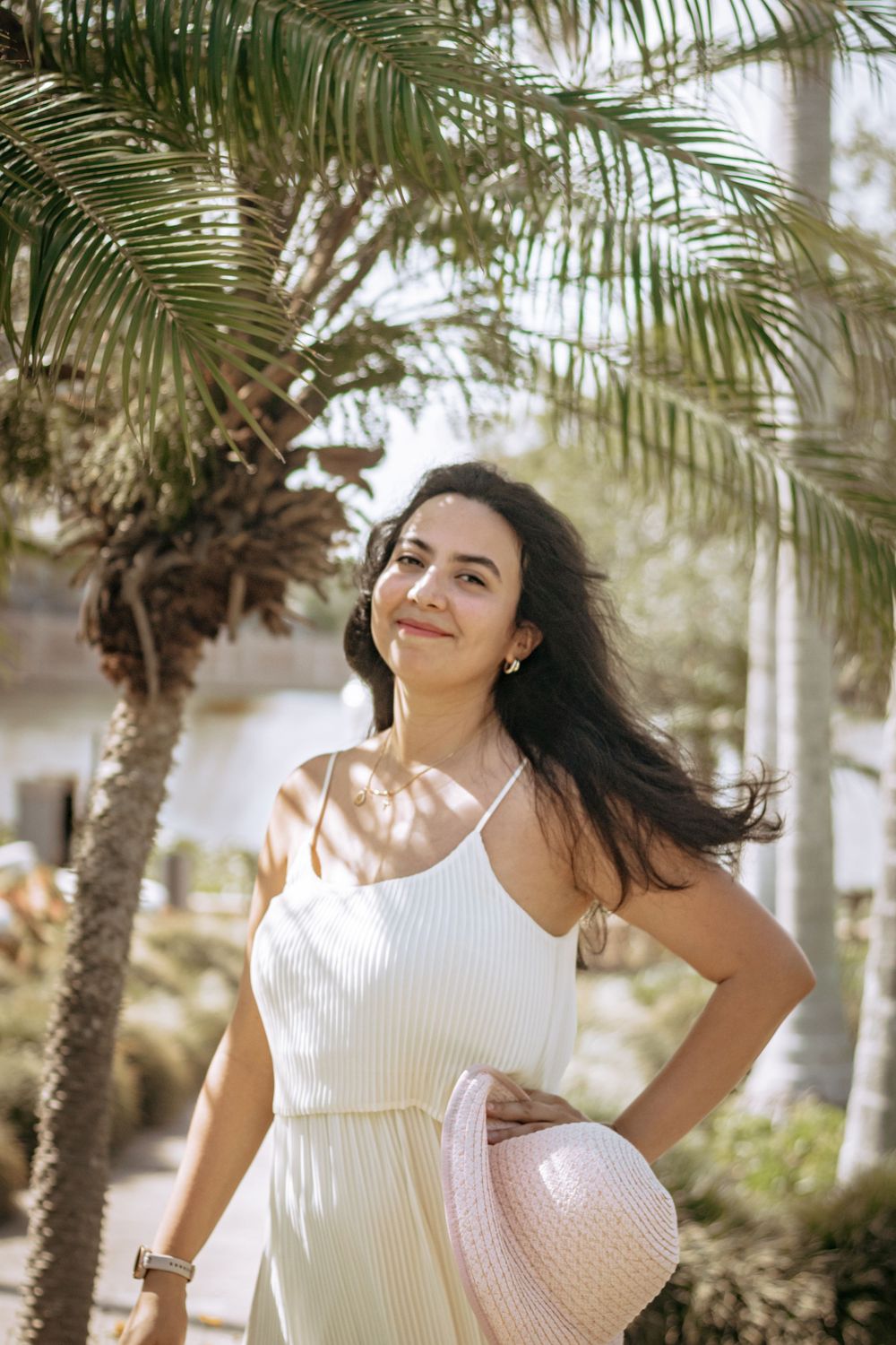 Young brunette woman in white sundress posing confidently under palm trees in sunlight.