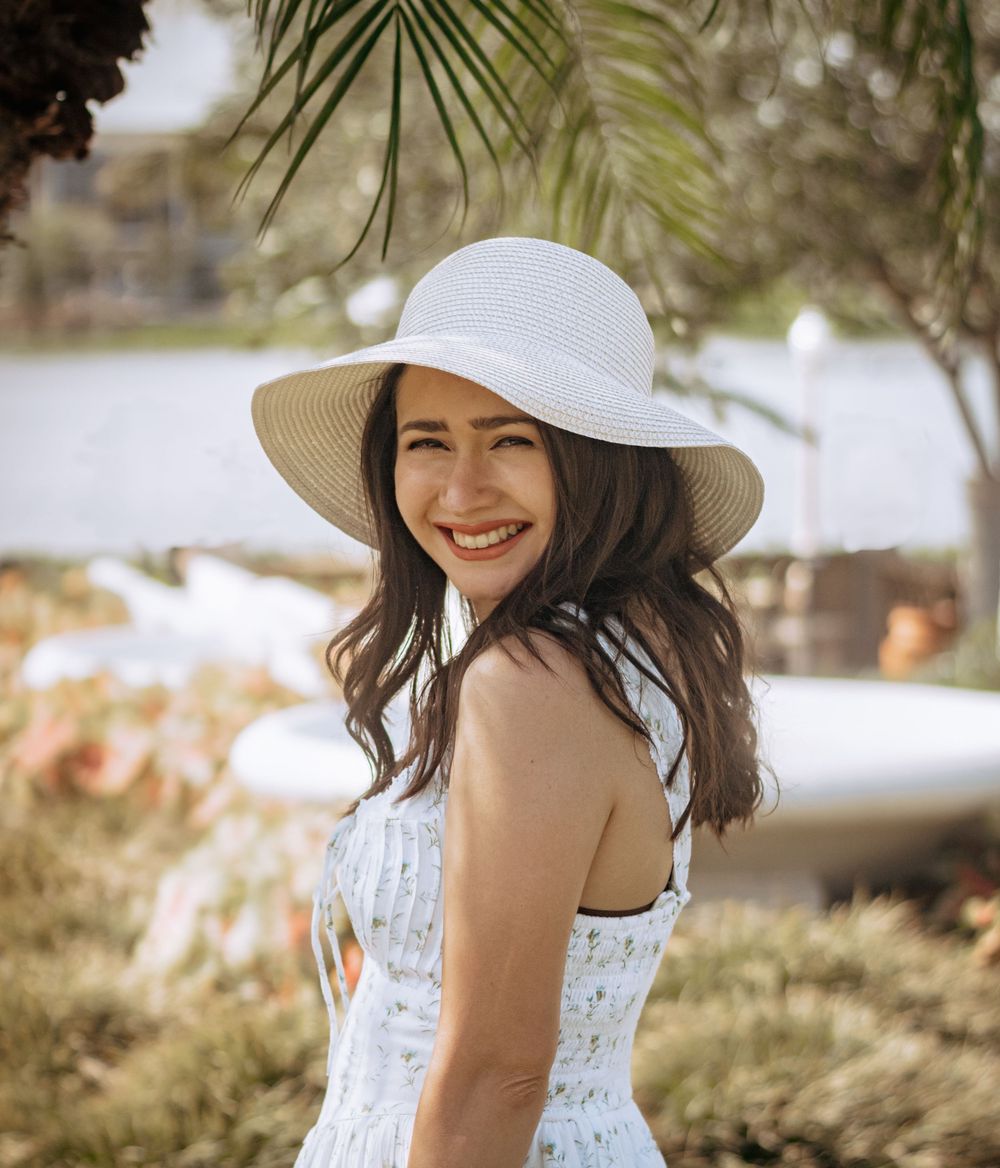 Cheerful brunette woman in white floral dress and sun hat smiling outdoors by a lake.