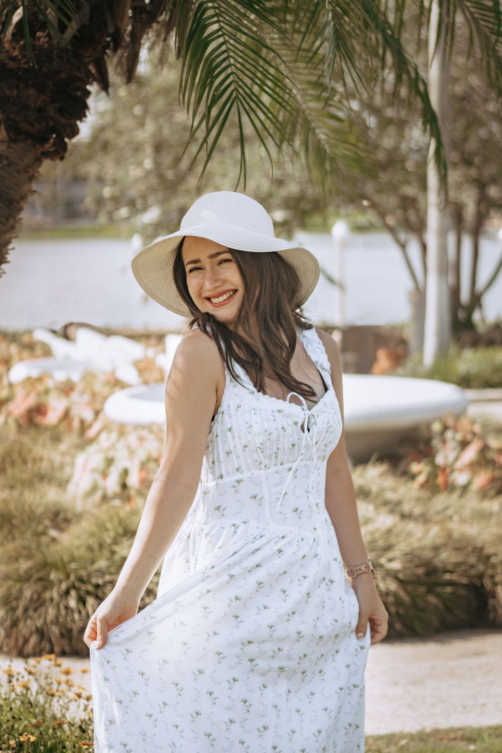 Happy woman in white floral midi dress and sun hat smiling under a palm tree.