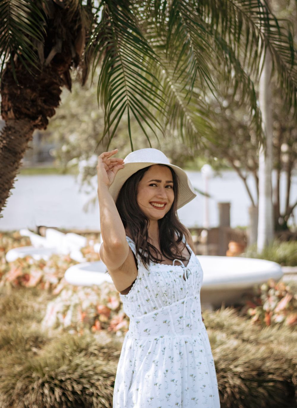 Joyful young woman in white dress holding sun hat up while standing near a palm tree.
