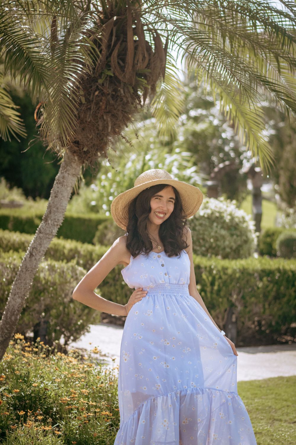 Smiling woman in light blue maxi dress and wide-brim straw hat posing in a lush tropical garden.