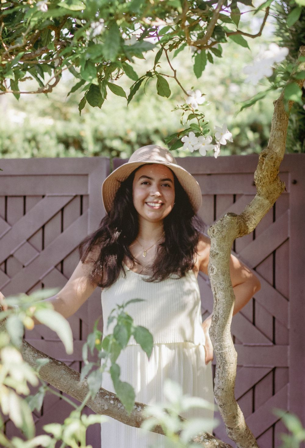 Smiling young woman in white dress and straw hat posing among flowering tree branches by a wooden fence.