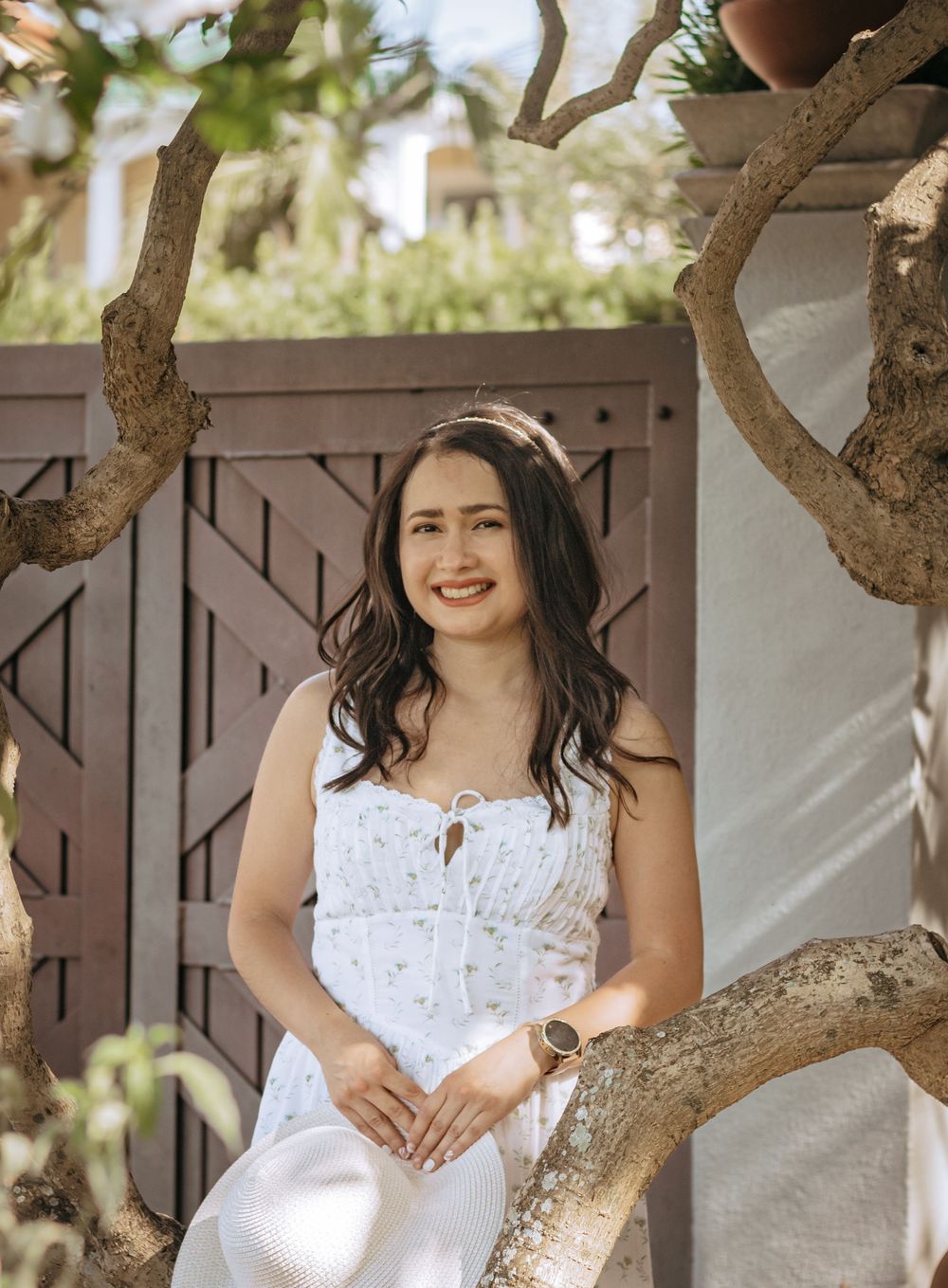 Young woman in white floral sundress sitting among tree branches smiling in a sunny garden setting.