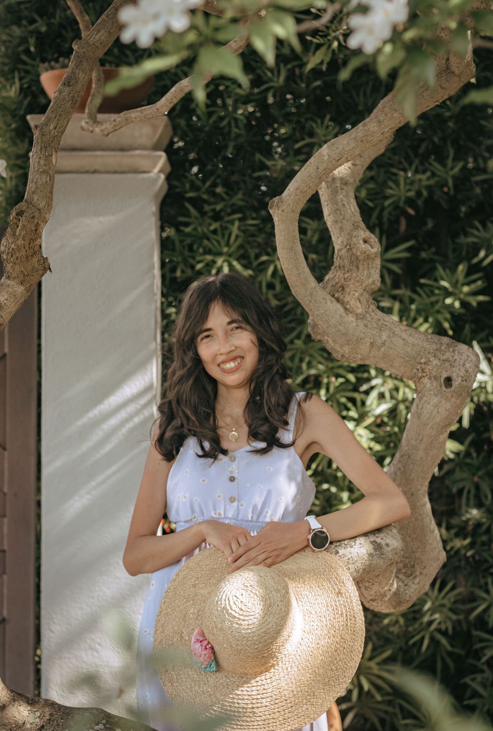 Happy young woman in blue top holding a straw hat while leaning against a tree in a garden.