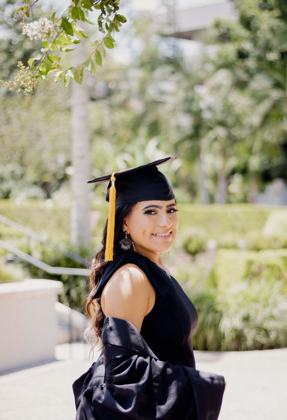 Graduate in black cap and gown with gold tassel smiling over shoulder in sunny outdoor setting.