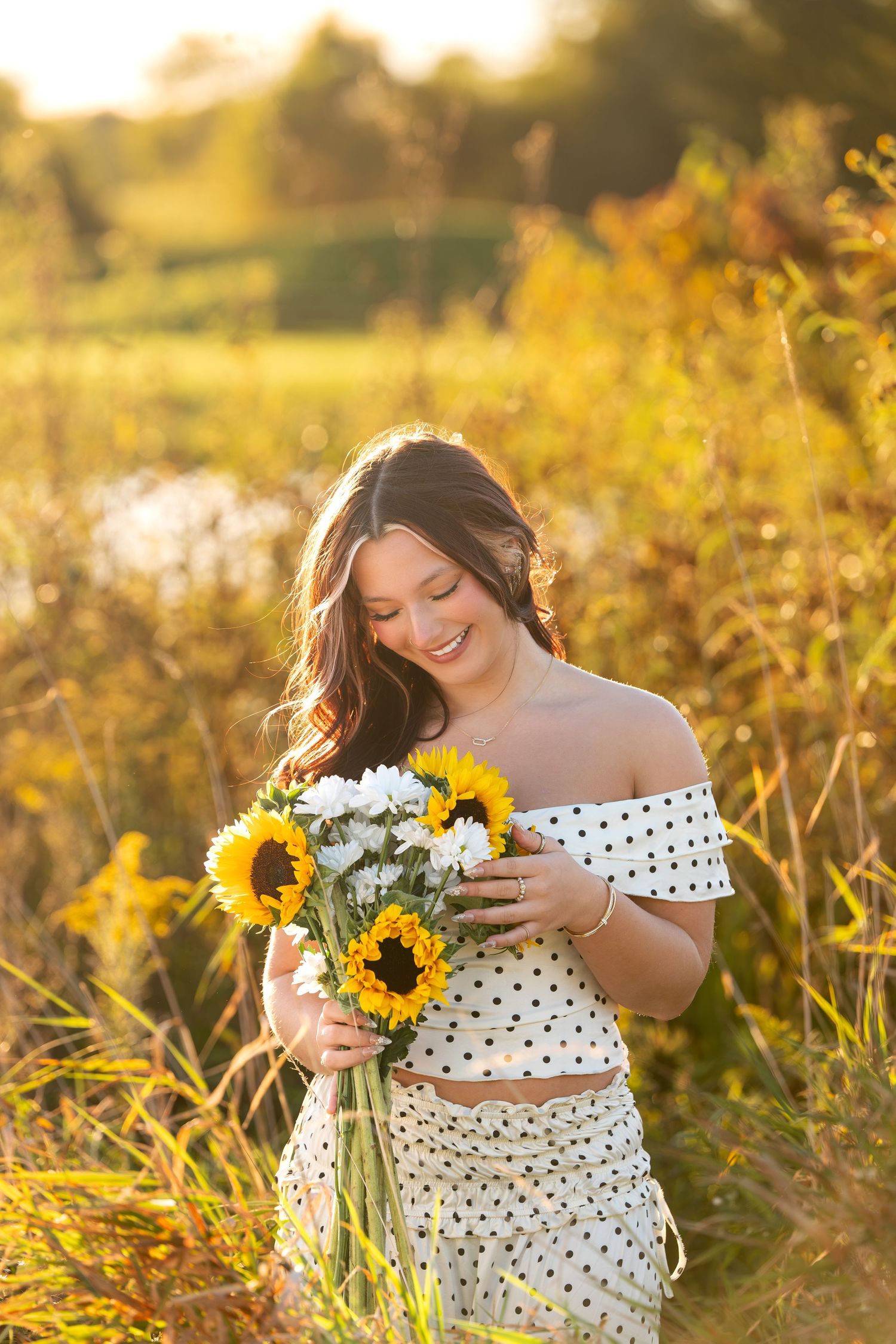 Smiling woman in a polka dot off-the-shoulder top and skirt holds sunflowers and white blooms in a golden field at sunset. youngstown senior