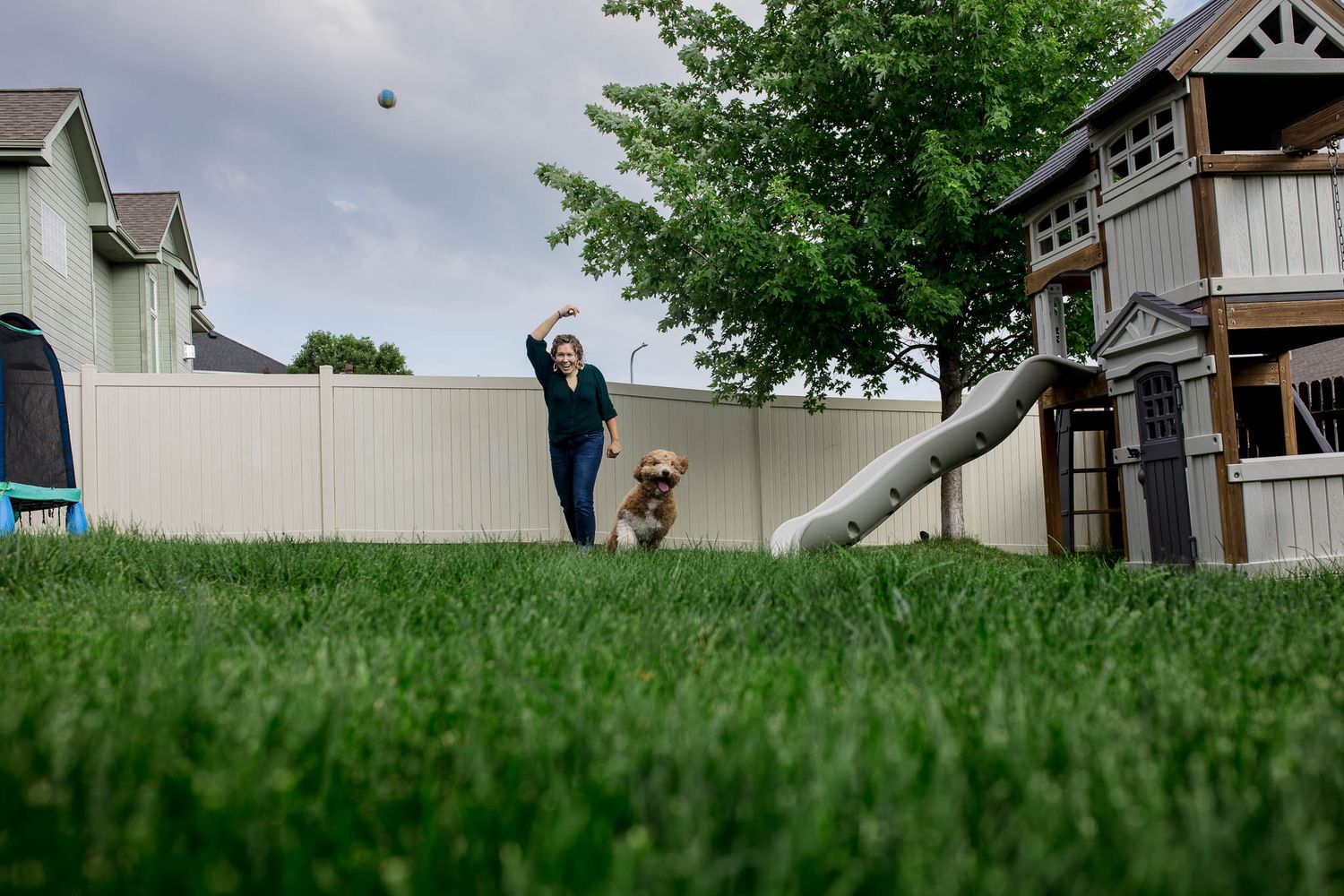 A backyard scene with a playground slide and someone playing fetch with a dog on lush green grass.