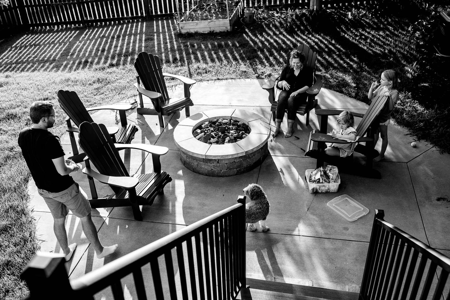 A backyard gathering around a fire pit shows people relaxing in chairs on a patio at dusk.