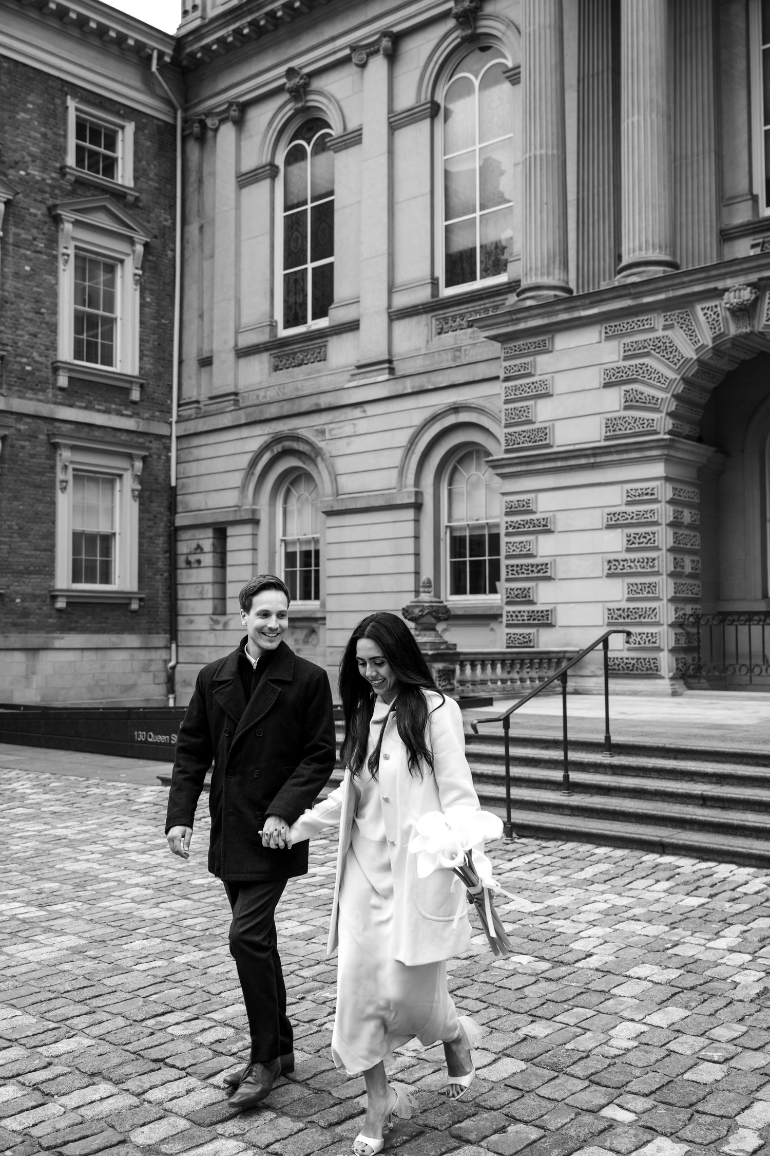 Two people walk hand in hand on a cobblestone path in front of a historic building with classical architecture.