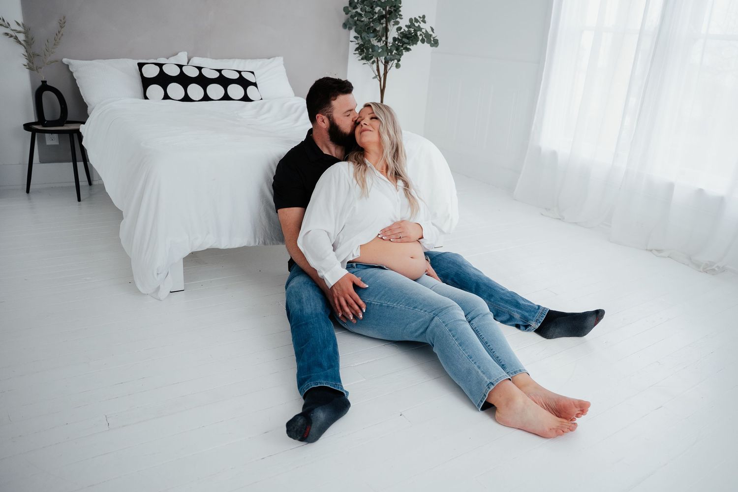 A couple sits together on white floor in bright minimalist bedroom with white bedding and plant decor.