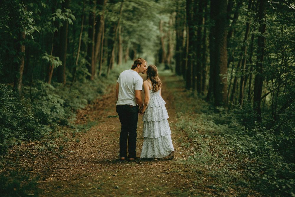 A couple in wedding attire share a romantic moment while standing on a wooded path surrounded by tall trees.