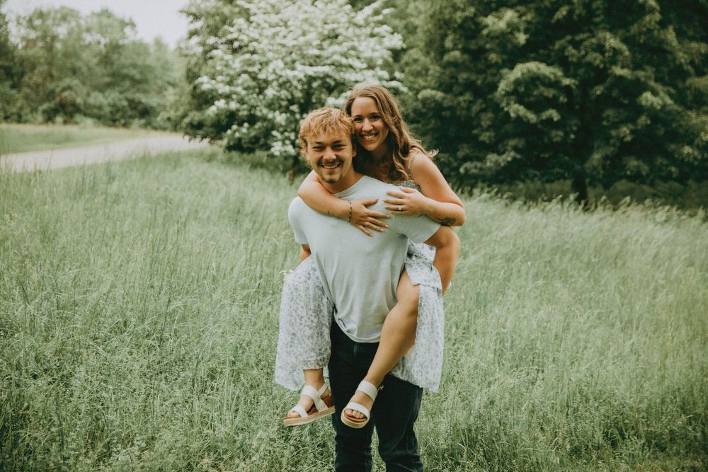 A couple shares a playful piggyback ride moment in a sunlit meadow surrounded by lush green grass and trees.