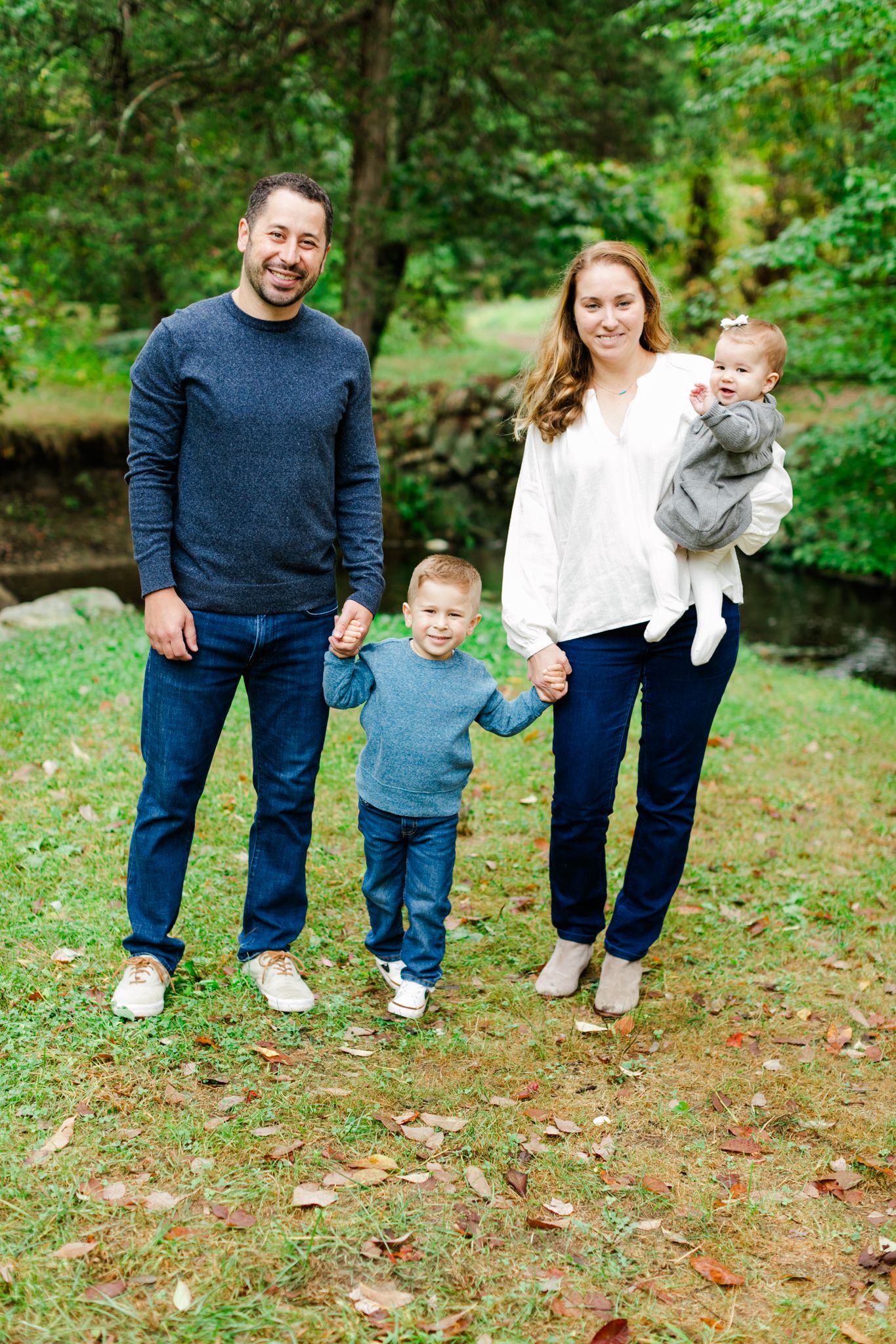 A family of four poses together outdoors in a wooded setting with fallen autumn leaves on the ground.