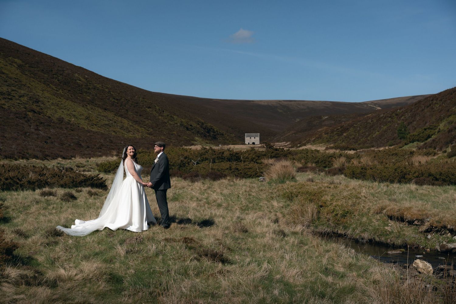 Newlywed couple stands in scenic moorland valley with rustic building in background on sunny day.