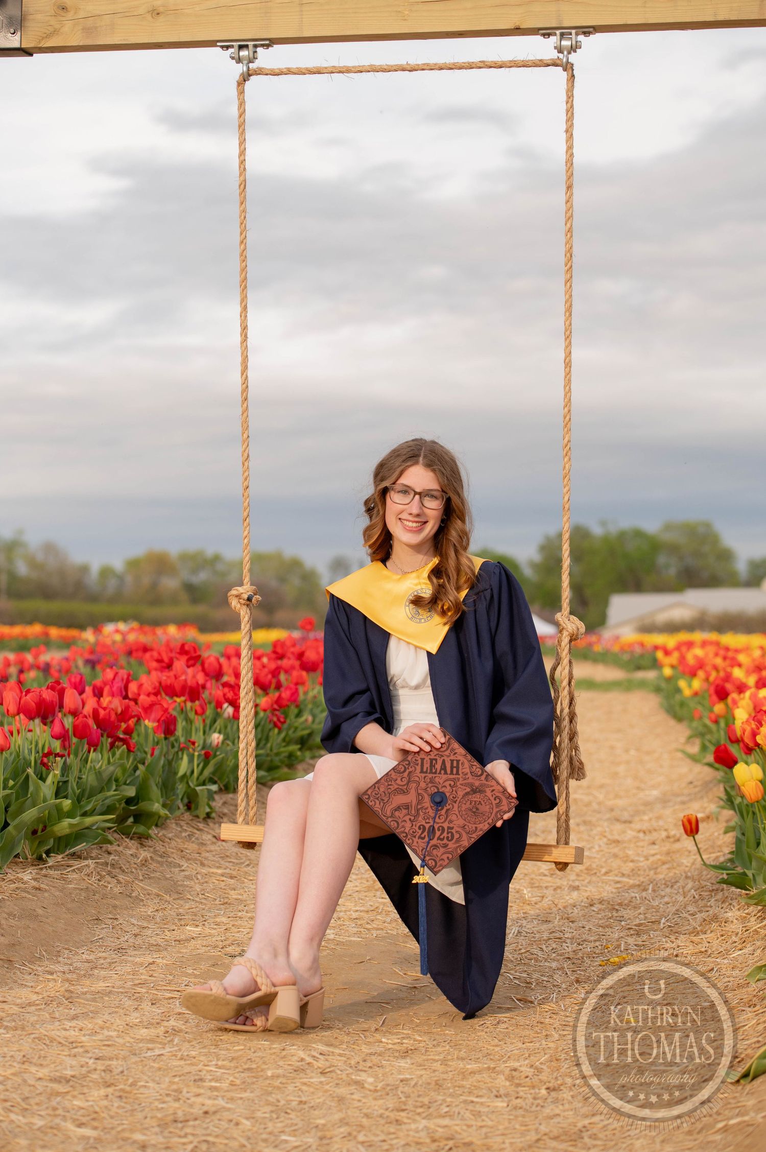 A graduate in cap and gown sits on a wooden swing surrounded by vibrant red tulip fields on a cloudy day.