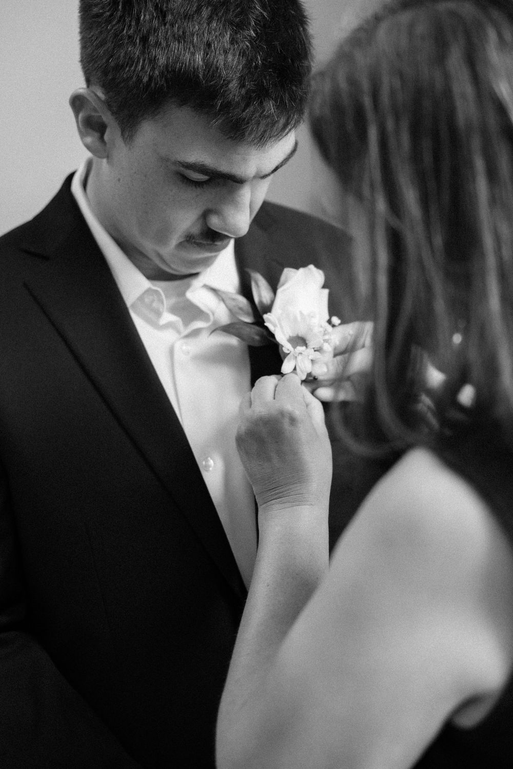 Black and white image of someone pinning a white floral boutonniere onto a formal dark suit jacket at a wedding.